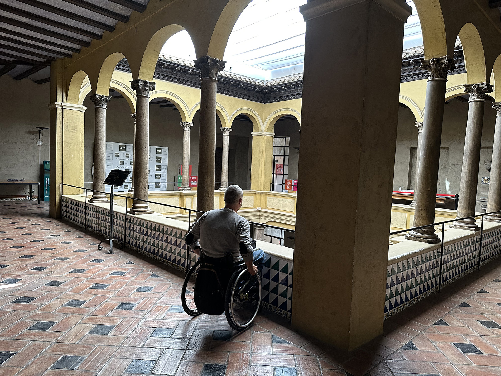 Hombre usuario de silla de ruedas en un patio interior con estilo arquitectónico tradicional y techo tragaluz.  El espacio está rodeado por una serie de columnas y arcos. La parte baja de la pared y el suelo es de cerámica con decoración geométrica.