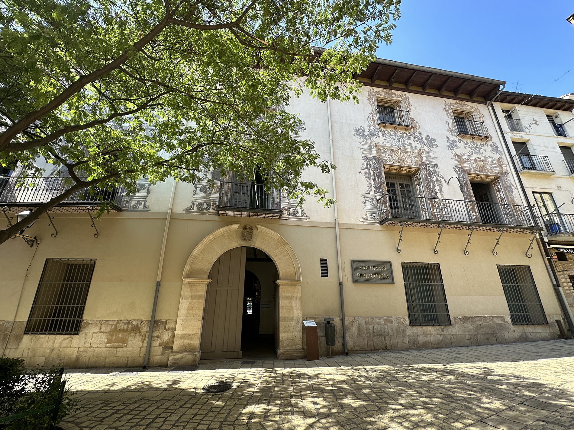 Fachada de un edificio histórico de tres plantas con balcones y decoraciones pintadas, acceso por un gran arco de piedra y placa que indica “Archivos Biblioteca”, parcialmente cubierta por las ramas de un árbol en una calle peatonal.