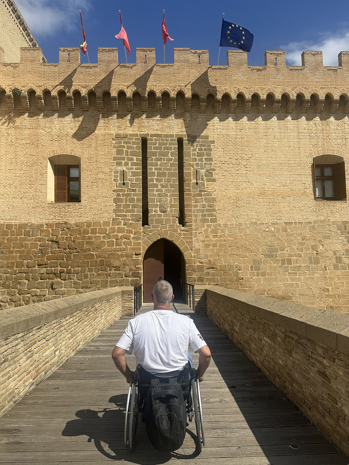 Persona usuaria de silla de ruedas cruzando un puente de madera hacia la entrada de un castillo de ladrillo con murallas y banderas en la parte superior.
