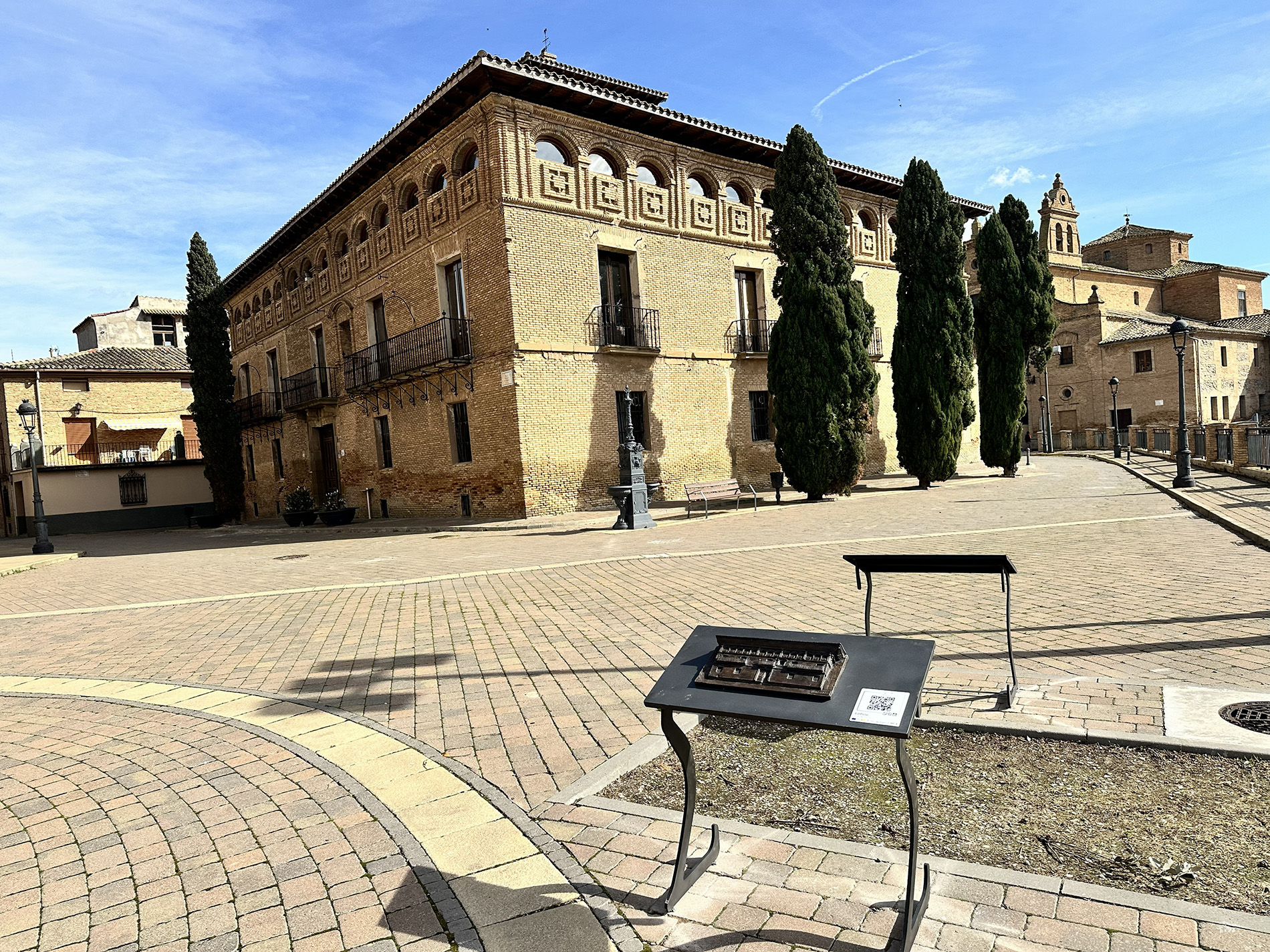 Plaza empedrada con un edificio histórico de ladrillo y balcones, cipreses alineados y un panel informativo en primer plano.